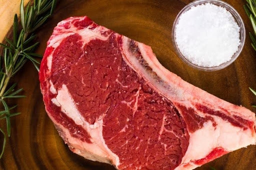 Raw bone-in ribeye steak with marbling, coarse salt, and rosemary on a wooden board, ready for how to cook ribeye steak guide.