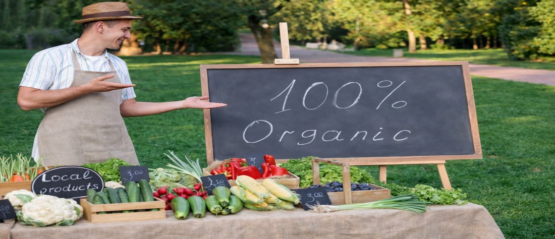 A farmer at a market stall displaying fresh produce with a “100% Organic” sign, illustrating what does organic mean in food labelling and local farming.
