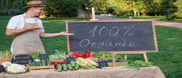 A farmer at a market stall displaying fresh produce with a “100% Organic” sign, illustrating what does organic mean in food labelling and local farming.